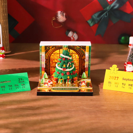 A Christmas-themed keepsake box displayed on a wooden table, surrounded by festive decorations including miniature Christmas trees, a Santa Claus figure, gift boxes, and holiday cards. The box features a clear acrylic top, a warm yellow holiday village scene printed on the inside, and a red-and-green argyle patterned base.